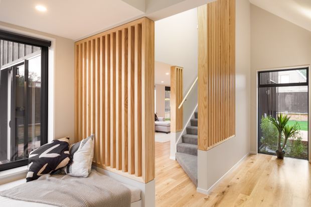 Bright contemporary residential interior showing a timber-lined stair partition and built-in window seat. Pale timber flooring, vertical timber battens, white walls, black-framed glazing, and a carpeted staircase create a warm, clean entry and circulation space with a glimpse toward a living area.