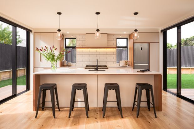 Bright contemporary kitchen with a large central island and four black bar stools, pale timber flooring, light-toned cabinetry, white tiled splashback, black-framed full-height glazing, and three pendant lights. The kitchen opens visually to a small lawn and fenced outdoor area.
