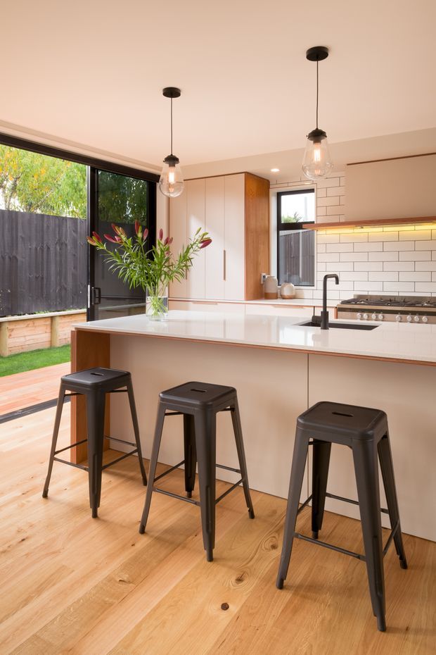 Bright contemporary kitchen with a large white island and seating for three, integrated cabinetry, timber accents, white subway-tile splashback, pendant lights, and black stools. Timber flooring and full-height glazed doors open toward a small outdoor area, creating an airy indoor-outdoor connection.