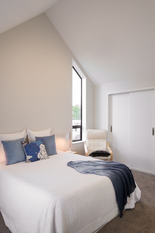 Bright, softly styled bedroom with a large white bed, white textured bedding, blue and patterned cushions, and a draped blue throw. The room has warm off-white walls, carpeted flooring, a tall narrow window, a white upholstered armchair, and white sliding wardrobe doors. The sloping ceiling creates a light, contemporary atmosphere.