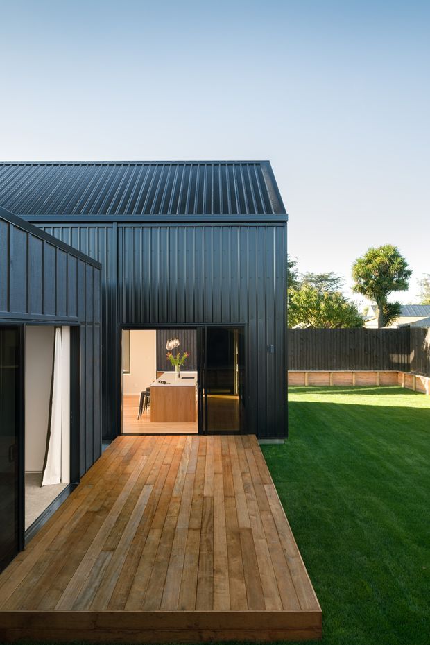 Exterior view of a compact dark-clad building with a steep corrugated metal roof, timber deck, glazed doors, and a neatly landscaped lawn. The deck leads toward an open doorway where a dining area is visible inside. The image has a clean contemporary character with strong vertical cladding and warm timber contrasting against the dark facade.