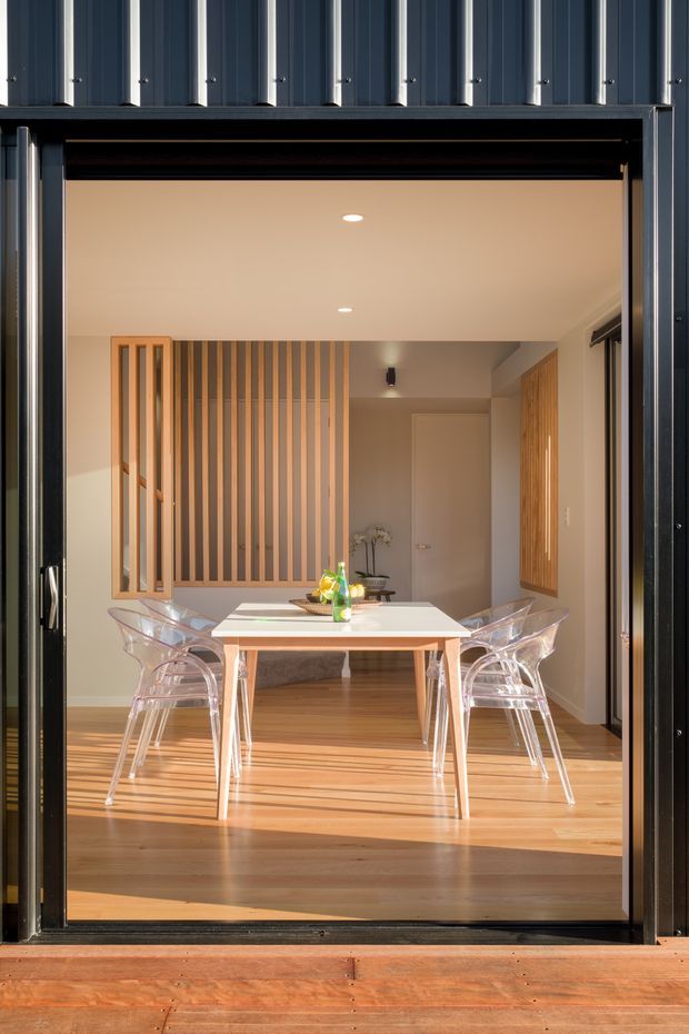 A bright dining area viewed through a large open black-framed glazed opening. The room features a light timber floor, white rectangular dining table, transparent acrylic chairs, warm timber vertical screening and cabinetry, and a simple white ceiling with recessed lights. The space opens onto a timber deck, with a dark corrugated metal exterior facade framing the view.