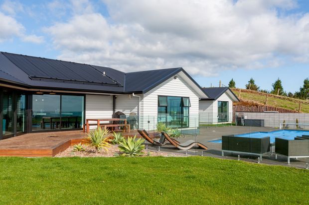 Contemporary single-storey residential home with white horizontal cladding, dark pitched metal roofs, extensive glazing, and a timber deck opening onto a landscaped lawn. Outdoor lounge furniture and a swimming pool occupy the foreground, with a planted sloping roof or earth-covered embankment visible behind the house.