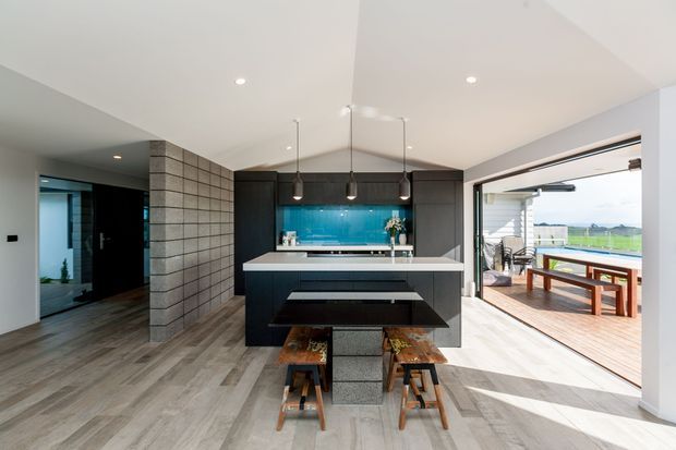Modern open-plan kitchen with a dark island and integrated seating, vivid turquoise glass splashback, recessed ceiling lights, and three pendant lights. The room features grey wood-look flooring, a textured grey block partition, and large retractable glazed doors opening to a timber deck with an outdoor dining table and landscaped views.