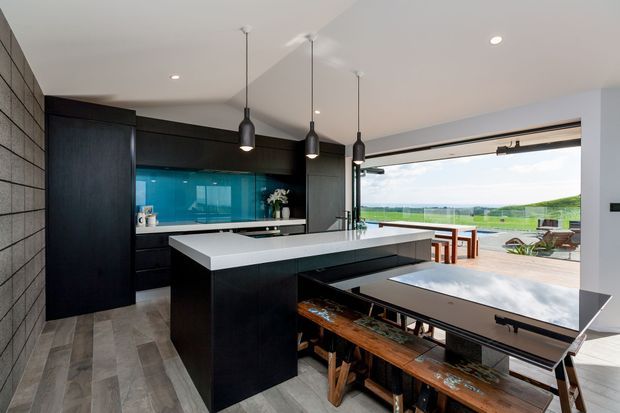 Contemporary open-plan kitchen and dining area with dark timber cabinetry, a large white island, turquoise glass splashback, and three black pendant lights. A long dining table occupies the foreground, while fully opened glazing connects the room to a spacious outdoor deck and expansive green landscape.