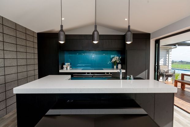 A contemporary kitchen centered on a large white stone island with integrated seating overhang. The kitchen features dark timber or laminate cabinetry, a vivid blue glass splashback, integrated appliances, three black pendant lights, and a textured grey tiled feature wall. Open glazing at right connects to a timber deck and bright outdoor view.
