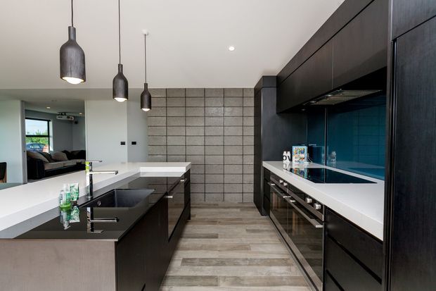 A contemporary galley-style kitchen with a large central island, dark timber or black cabinetry, white stone-look benchtops, integrated appliances, and a blue tiled splashback. A grey block-pattern feature wall and pendant lights add texture, while an open living area is visible beyond the kitchen.