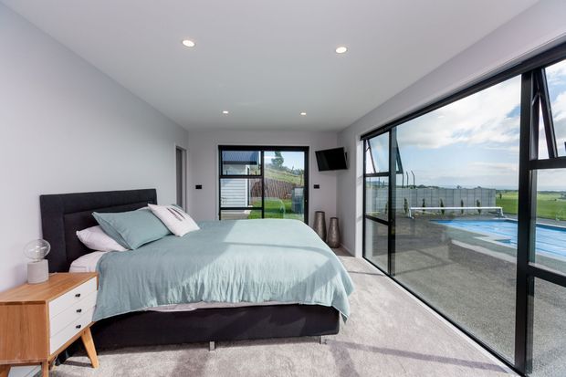 Bright contemporary bedroom with a king-size bed, pale blue bedding, dark upholstered headboard, timber bedside table, recessed ceiling lights, and expansive black-framed floor-to-ceiling glazing. The glazing opens to a terrace and swimming pool, with rural views beyond.
