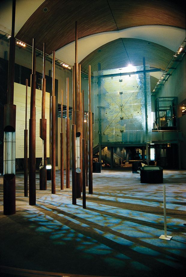 A large dramatic interior space resembling a museum, gallery, or cultural exhibition hall. Tall slender copper-toned vertical elements form an installation in the foreground, while a monumental translucent blue-green glazed or mesh-like volume and stair structure occupy the background. The space has a high curved ribbed timber ceiling, exposed structural and display elements, polished hard flooring, and theatrical track lighting. Strong blue and amber illumination creates patterned shadows across the floor.