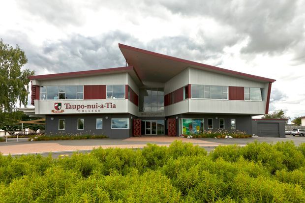 Exterior view of a two-storey contemporary school building with a distinctive asymmetrical, overhanging roof and red-edged roofline. The facade combines grey lower walls, light corrugated-looking upper cladding, extensive horizontal windows, red vertical accent panels, and a glazed recessed central entrance. Landscaping and a paved frontage sit in the foreground.