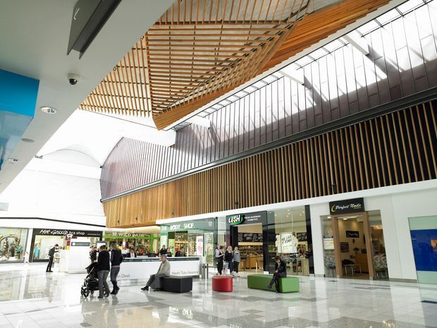 Bright contemporary indoor shopping centre atrium with polished tiled flooring, retail storefronts, a high glazed roof, and extensive warm timber slatted screens and suspended timber ceiling structures. The open public space includes seating, information desks, shoppers, and multiple shops.