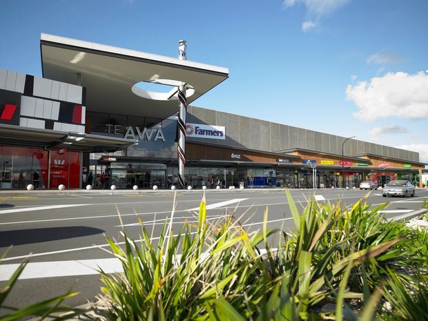 Exterior view of a contemporary suburban shopping centre, with a broad commercial frontage containing retail shopfronts, Farmers and BNZ signage, a projecting flat canopy, and a large paved forecourt with road markings and planted flax-like landscaping in the foreground.