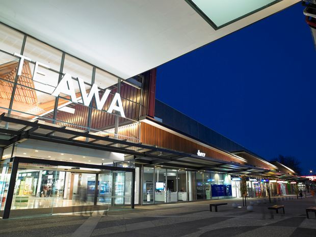 Dusk exterior view of a contemporary commercial retail and hospitality complex, featuring a broad projecting canopy, glazed shopfronts, illuminated signage, timber-look vertical cladding, metal framing, and a paved pedestrian forecourt. The large white 'TAWA' lettering is prominent on the upper facade.