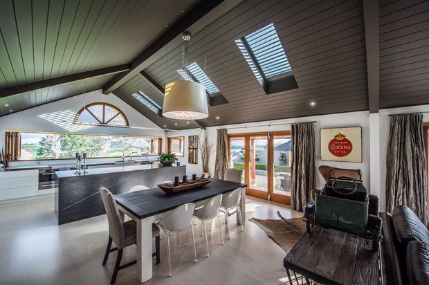 A spacious open-plan kitchen and dining area with a dramatic vaulted timber-lined ceiling, exposed dark beams, multiple skylights, and clerestory-style glazing. The kitchen features sleek white cabinetry, a dark stone island and benchtop, and stainless-steel appliances. A large dark timber dining table sits centrally beneath an oversized pendant light, with glazed timber-framed doors opening to an outdoor view. The room also includes dark leather seating, patterned curtains, and eclectic decorative elements.