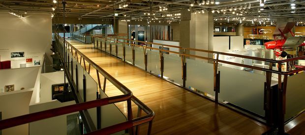 A wide interior view of a multi-level commercial or exhibition space, centered on a polished timber walkway and mezzanine with glass balustrades and dark metal framing. The open void reveals lower-level areas, while track and string lighting illuminate exposed ceiling services. Display installations and signage are visible in the surrounding gallery-like environment.