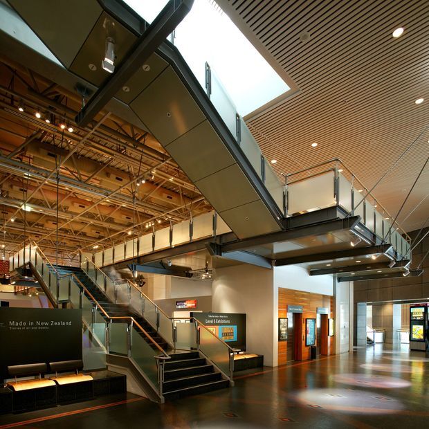 A large contemporary public interior with a dramatic multi-level circulation layout, exposed structural and mechanical services, extensive staircases and mezzanine walkways. The space combines a warm slatted timber ceiling with steel framing, glass balustrades, polished dark flooring, lift or service doors, and illuminated signage. The architecture reads as a civic, cultural, or institutional building lobby rather than a residential room.