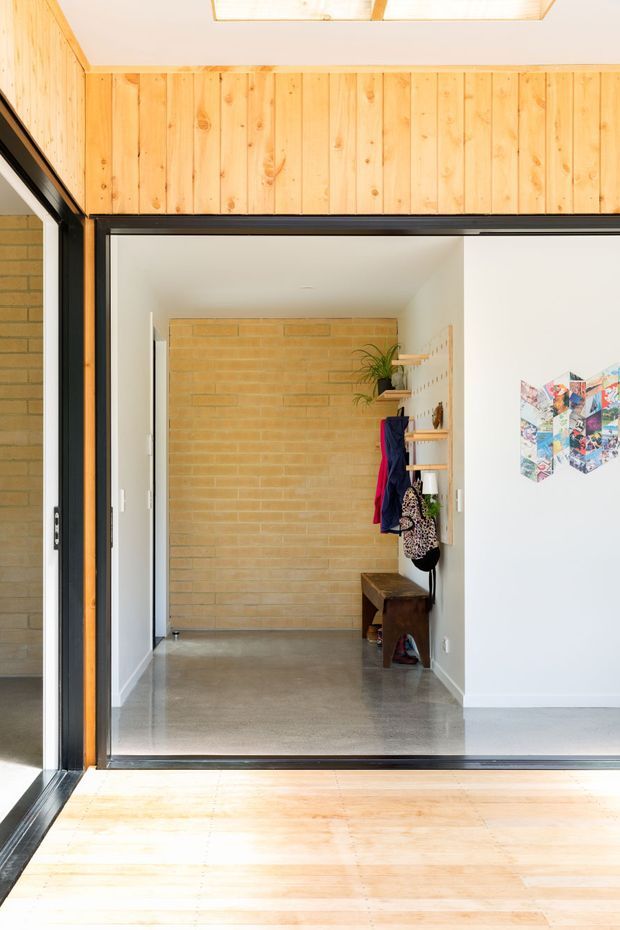 Bright architectural view into a compact entry hall through a large black-framed glazed opening. The space features warm vertical timber wall panelling, a pale brick feature wall, polished grey concrete flooring, white walls, built-in open shelving with coats and a plant, and a simple timber bench. A skylight or roof window is visible above, with timber flooring in the foreground.
