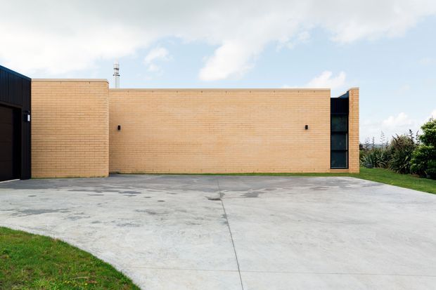 A low, modern brick building viewed from the front across a broad concrete driveway. The facade is composed of long warm tan brick walls, a dark recessed entrance or side volume at left, and a narrow full-height glazed opening near the right edge. Minimal exterior detailing and a flat roofline create a clean, understated architectural composition, with lawn and planting bordering the driveway.