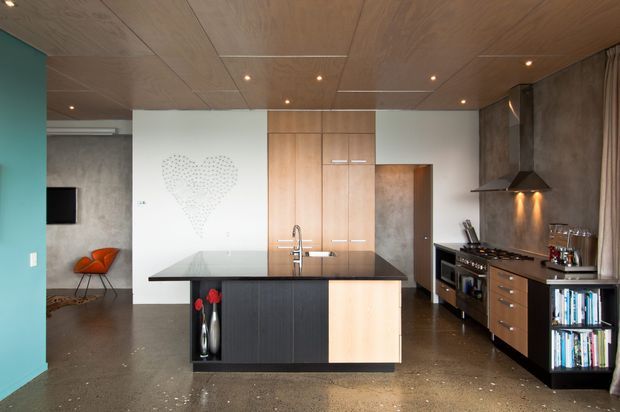Open-plan contemporary kitchen with a central island, dark stone benchtop, timber and dark cabinetry, integrated appliances, stainless-steel rangehood, and a timber-paneled ceiling. The space opens toward a living area with a turquoise accent wall and concrete flooring.