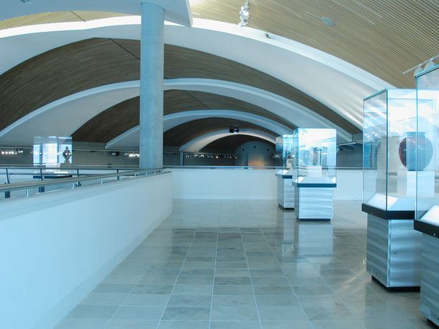 A spacious, modern museum or gallery interior with a dramatic barrel-vaulted ceiling formed by repeated curved ribs and warm wood slats. The bright white structure includes large cylindrical columns, extensive open circulation space, polished light stone or ceramic tile flooring, stainless-steel and glass display cases, and glass balustrades. The setting is clean, airy, and highly architectural, with cool blue-white lighting.