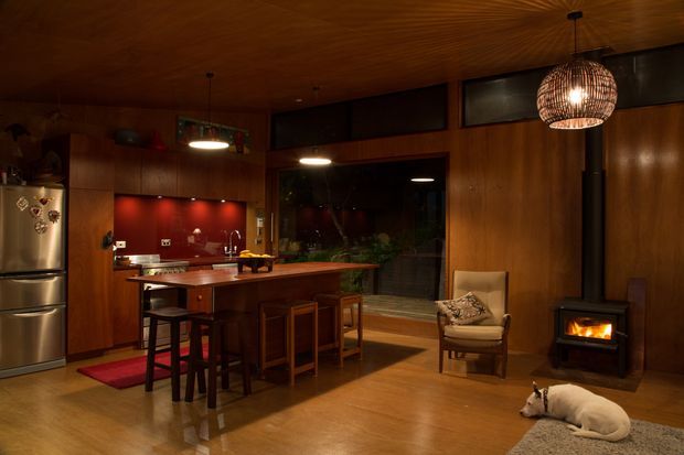 Warm, compact open-plan kitchen and living area with extensive dark timber wall and ceiling panelling. The kitchen features a central timber island with bar stools, dark cabinetry, a red backsplash, and a stainless-steel refrigerator. The adjoining living zone includes a freestanding wood-burning stove, a woven pendant light, and a lounge chair, with large glazed openings providing views into a planted outdoor area.