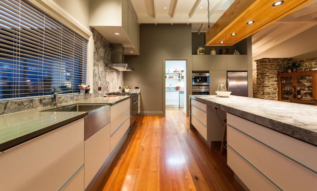 Contemporary galley-style kitchen with a central island, extensive pale cabinetry, dark stone benchtops and splashback, stainless-steel appliances, and warm polished timber flooring. A large window with timber blinds, exposed timber ceiling beams with recessed lighting, and a rustic exposed-brick feature wall add texture and contrast.