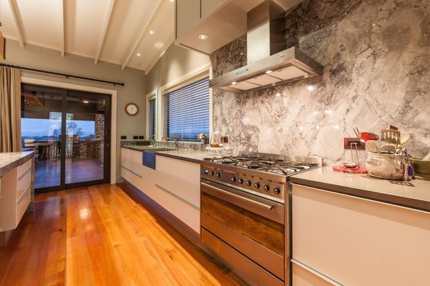 A refined contemporary kitchen with a long galley-style layout, extensive light cabinetry, a large stainless-steel range cooker and integrated rangehood, and a dramatic full-height veined stone splashback. Warm timber flooring contrasts with the pale cabinetry, while a raked white ceiling with exposed beams and clerestory-style glazing adds height and natural light. Sliding glass doors connect the kitchen to an adjoining outdoor or living area.