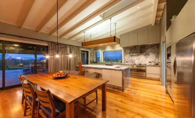 Warm, expansive open-plan kitchen and dining area with a large timber dining table, upholstered timber dining chairs, central stone island, extensive built-in cabinetry, stainless-steel appliances, and a textured stone splashback. Exposed light timber ceiling beams and pendant lighting add character, while full-height glazing opens toward an outdoor view.
