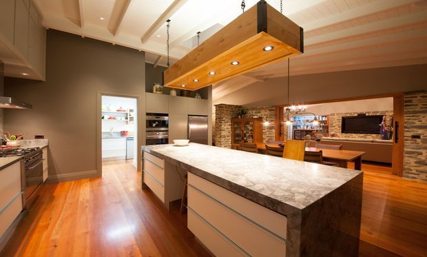 Open-plan kitchen with a large stone-topped island, integrated stainless-steel appliances, warm timber flooring, and a substantial timber pendant light. The kitchen opens to a dining and living area with exposed brick and stone feature walls, timber beams, and a wall-mounted television.