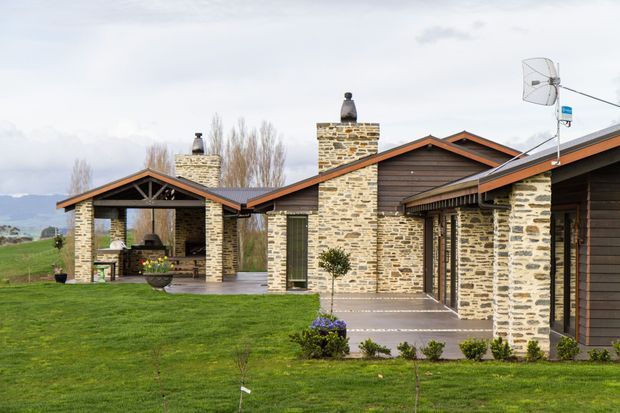 Single-storey residential house set in a rural landscape, featuring extensive irregular natural-stone masonry walls and chimneys, dark horizontal timber cladding, and multiple pitched gabled roof forms with wide eaves. A covered outdoor entertaining area with a fireplace is visible at left, while paved paths and manicured lawn surround the building. The design has a rustic contemporary lodge or farmhouse character.