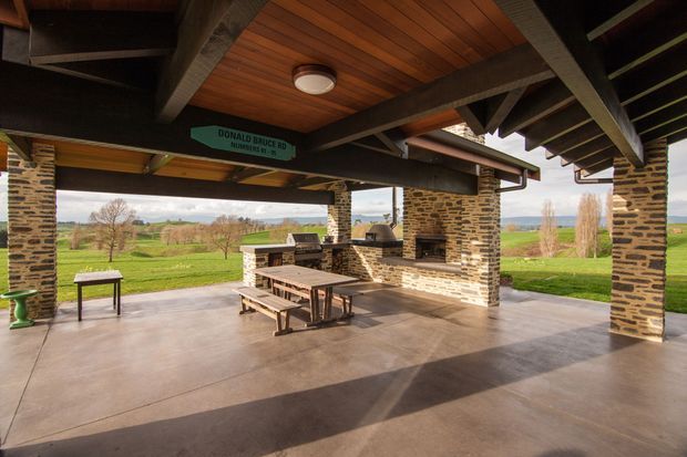 A spacious covered outdoor entertaining area with a dark timber-beamed ceiling, warm timber soffit, stone-clad columns, polished concrete flooring, and a built-in outdoor barbecue bench. A large timber dining table and benches sit beneath the roof, opening onto expansive rural farmland views.
