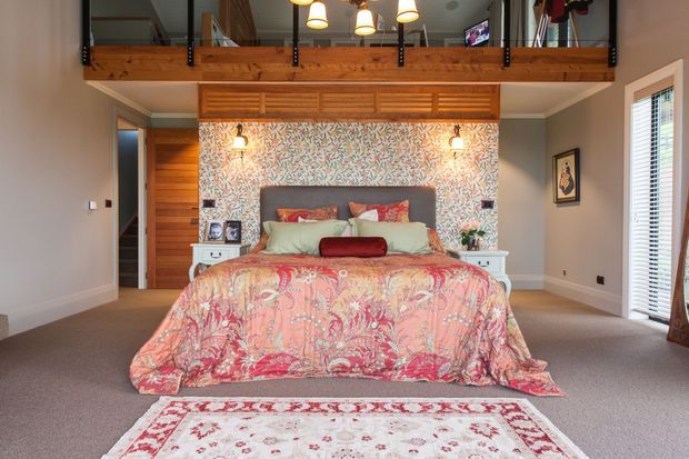 A warm, richly layered bedroom with a large bed dressed in a coral and pink patterned quilt, pale green pillows, and a dark upholstered headboard. The room features floral patterned wallpaper, timber wall panelling and an overhead timber mezzanine or loft with black metal railings. Carpeted flooring, bedside tables, framed artwork, wall sconces, and pendant lighting complete the inviting interior.
