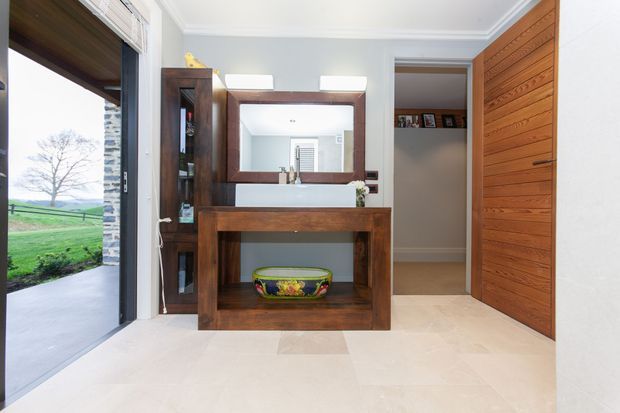 Bright, refined residential entryway featuring a dark timber console with integrated shelving, a large framed mirror, and a warm horizontal timber-panelled door. Pale stone-look flooring, light grey walls, white decorative ceiling trim, and a glazed opening to the landscaped exterior create an inviting transition space.