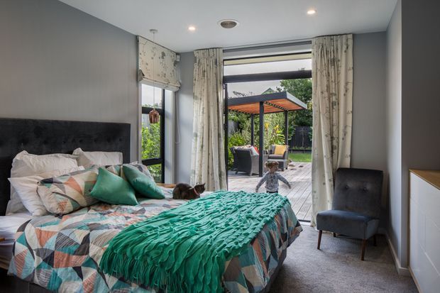 A bright contemporary bedroom with a large upholstered dark headboard, patterned geometric bedding, teal cushions and throw, and full-height glazed doors opening to a paved outdoor patio. Floral curtains frame the opening, while a dark accent armchair and white built-in cabinetry complete the room. An outdoor pergola seating area is visible beyond the glazing.