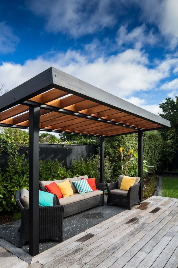 A contemporary outdoor covered lounge structure set within a leafy garden. The pergola features a broad flat roof with exposed warm timber slats beneath, dark structural posts and beams, and a weathered timber deck. Wicker seating with colourful cushions creates a relaxed entertaining area, enclosed by planting and a dark fence beneath a partly cloudy blue sky.