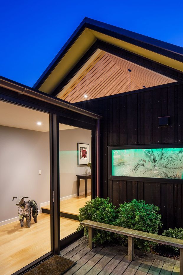 A dusk exterior view of a contemporary residential entry courtyard. Black vertical timber cladding frames a covered porch with a warm timber-lined soffit, recessed lighting, and dark-framed glazed entry doors. A timber deck, built-in planter, shrubs, and a narrow illuminated decorative window feature sit beside the entrance. The open doorway reveals a softly lit interior hallway or entry space with pale timber flooring and a console table.