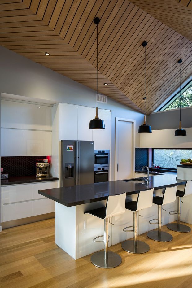 A contemporary kitchen with white flat-panel cabinetry, a large dark island with an integrated sink and seating for four, stainless-steel appliances, and a dark hexagonal-tile splashback. The room features a warm timber-lined sloping ceiling with recessed downlights and three black pendant lights, timber flooring, white bar stools, and a long horizontal window providing natural light.