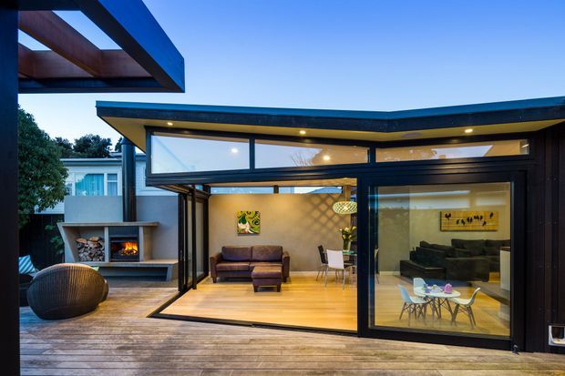 A dusk view from a timber deck into a contemporary pavilion-style living area with extensive black-framed glazing and sliding glass doors. The open-plan room contains a leather sofa, lounge chair, dining table, pendant light, artwork and a built-in fireplace set into a concrete outdoor wall. A flat, projecting roof with clerestory glazing and exposed timber elements creates a strong indoor–outdoor connection.