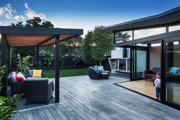A contemporary residential outdoor entertaining area with a large weathered timber deck, covered pergola, outdoor seating, and a central open courtyard. The adjacent pavilion has extensive dark-framed glazing, a high sloping roofline, timber flooring, and a built-in fireplace visible inside. Dense garden planting surrounds the space.