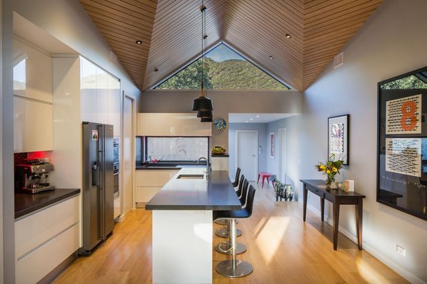 Bright contemporary kitchen and dining area with a long white island, integrated stainless-steel appliances, dark stone benchtops, timber flooring, and a dramatic pitched timber-lined ceiling. A large triangular clerestory window brings in natural light and frames a leafy hillside view. The island includes seating for four, while a slim console table and framed artwork occupy the adjacent wall.