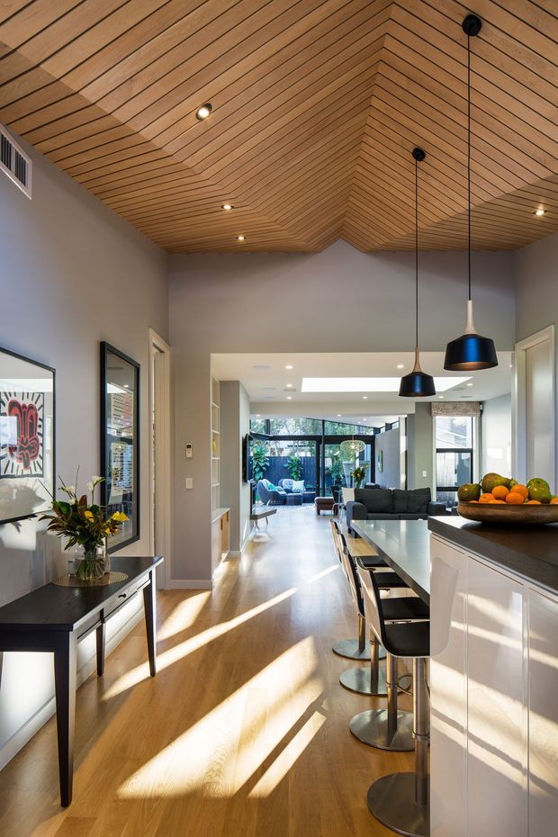 Bright open-plan kitchen and living area with a long white island and dark stone benchtop, integrated bar seating, timber flooring, and a dramatic warm timber-lined vaulted ceiling with recessed and pendant lighting. The space opens toward a glazed living area and courtyard, with framed artwork and a console along the side wall.