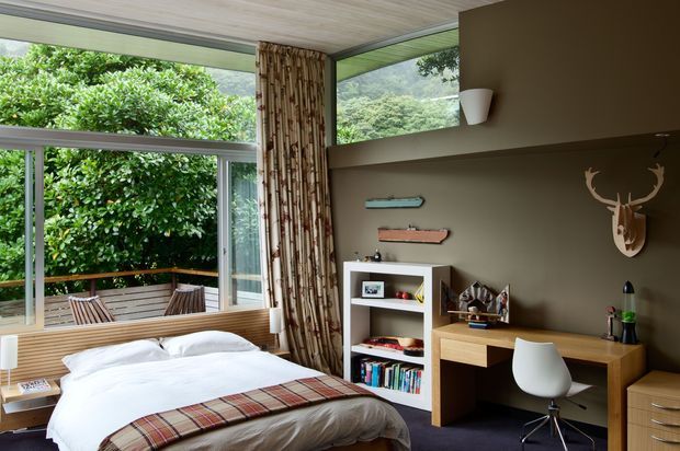 A modern bedroom with a low platform bed dressed in white bedding and a plaid throw, positioned beside expansive corner glazing that opens toward a leafy outdoor deck. The room features a warm wood ceiling, wood-framed glazing, a textured vertical timber feature screen, built-in bedside ledge, open shelving, compact timber desk, white molded chair, drawers, wall-mounted shelves, and a sculptural deer-head wall decoration. The palette is warm and earthy with strong indoor–outdoor connection.