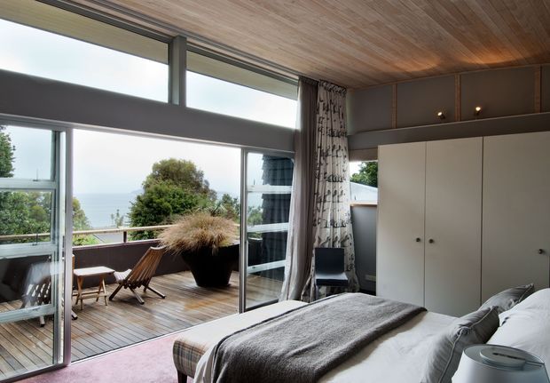 A warm, compact bedroom opening onto a timber deck through extensive floor-to-ceiling glazing and sliding doors. The room has a built-in white wardrobe, patterned curtains, a timber-lined ceiling, and a neatly made bed. Outdoor seating and lush vegetation are visible on the deck, with a distant water view beyond.