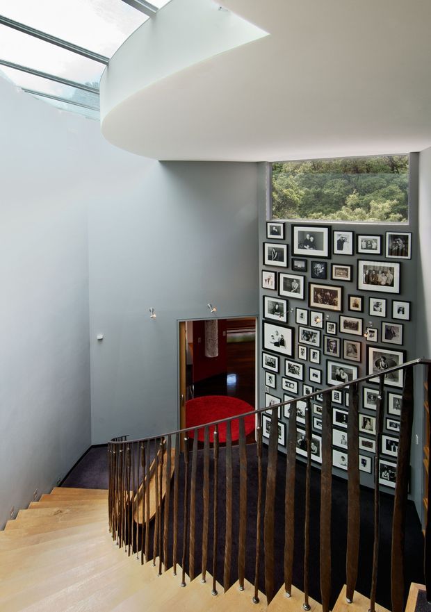 A contemporary interior stairway viewed from an upper landing, with pale timber stairs, dark slender metal balustrades, and a sweeping curved white ceiling above. A large skylight brings daylight into the tall stairwell. The wall beside the stairs is densely arranged with framed black-and-white and colour photographs, while a red upholstered chair and darker adjoining room are visible below.