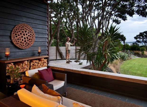 A covered outdoor lounge terrace beside a dark horizontal-clad dwelling, furnished with outdoor seating and cushions. A built-in timber feature wall includes a firewood storage niche and decorative wall art, with candles adding warmth. The terrace overlooks a landscaped garden with mature trees, sculptural palms, lawn, and a distant coastal or water view.