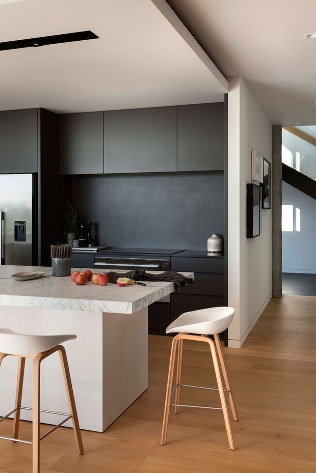 A contemporary open-plan kitchen viewed from the adjoining living area, with a dark matte built-in cabinetry wall, integrated stainless-steel refrigerator and appliances, dark splashback, and a pale stone island with waterfall edge. Two light upholstered timber bar stools sit at the island. Warm timber flooring, white walls, framed artwork, and a high sloping ceiling create a refined, minimalist interior. A hallway and part of another bright room are visible beyond the kitchen.