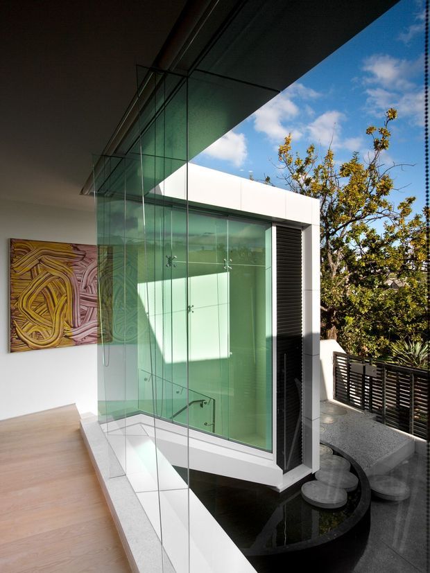 A contemporary interior overlooking a glazed, double-height stairwell or void. Floor-to-ceiling glass balustrades and a large green-tinted glass enclosure frame a sculptural staircase, with pale timber flooring, white walls, dark stone or tile flooring, and a black louvred screen. A small water feature with stepping stones sits beside the glazing, while an abstract artwork adds colour to the wall. The space is brightly lit by strong daylight and opens visually to an outdoor terrace and trees.