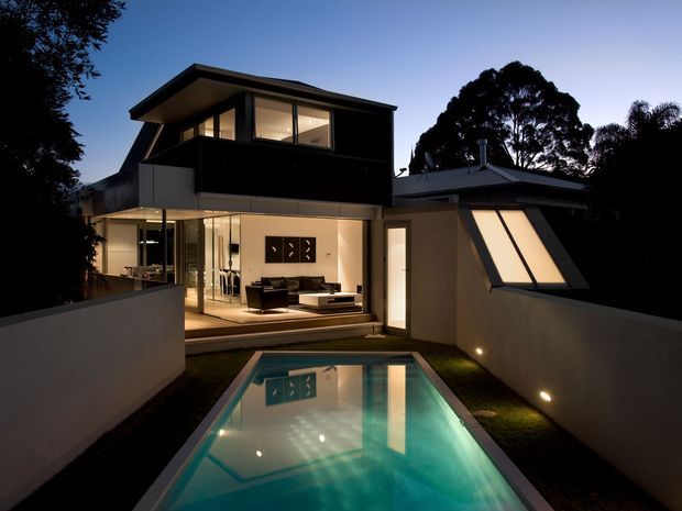Twilight exterior view of a contemporary multi-level residence with strong geometric forms, extensive floor-to-ceiling glazing, and a small illuminated swimming pool in the foreground. The home has dark upper cladding, pale rendered walls, a low-pitched roof, and a projecting angled window element. A brightly lit living room is visible through the glazing.