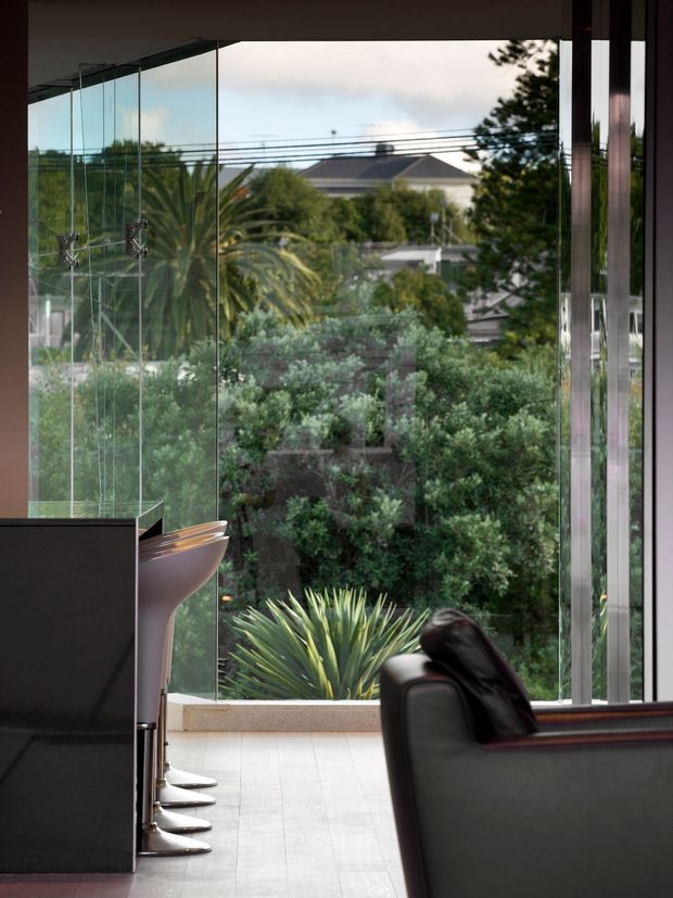 A contemporary interior with a dark built-in counter or partition at left, sculptural pedestal-like fixtures, and a black leather lounge chair in the foreground. Full-height glass walls open views toward a densely planted garden and neighbouring homes. The space has a restrained, refined palette of black, grey, timber-brown and muted green, with strong indoor-outdoor visual connection.