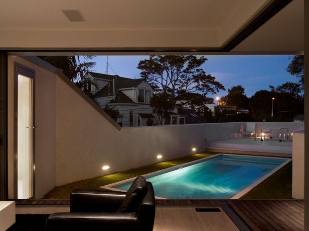A dusk view from a covered interior space onto a compact illuminated swimming pool courtyard. The pool is bordered by timber decking, a narrow grass strip, rendered privacy walls and subtle recessed wall lighting. A black leather lounge chair occupies the foreground, while a neighbouring traditional timber-clad house and trees are visible beyond the courtyard wall.